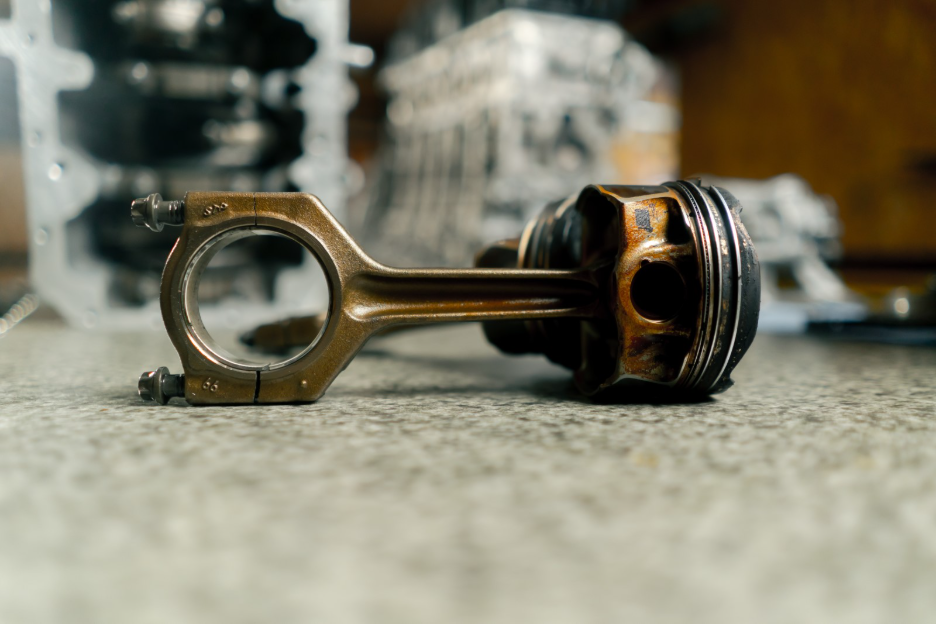 Close-up of a used engine piston and connecting rod resting on a workbench, with engine components blurred in the background